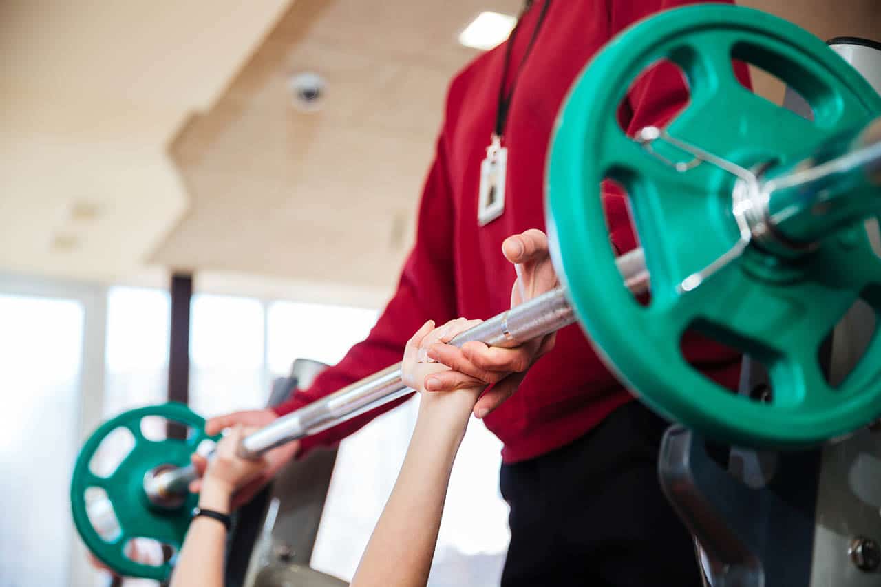 Closeup of hands of young sportswoman training with fitness instructor using barbell in gym
