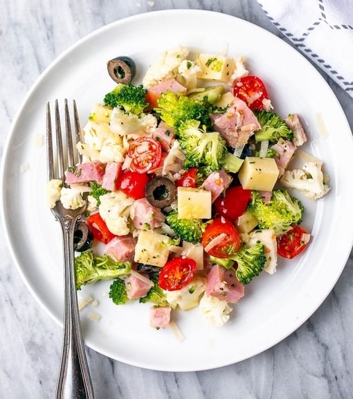 Brokolicovo-květákový salát se šunkou, sýrem, olivami a rajčátky na bílém kulatém talíři